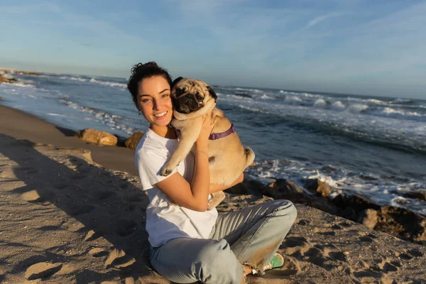 Positive young woman with curly hair holding pug dog on beach near sea in Barcelona — Stock Photo