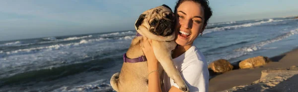 Cheerful young woman with curly hair holding pug dog on beach near sea in Barcelona, banner — Stockfoto