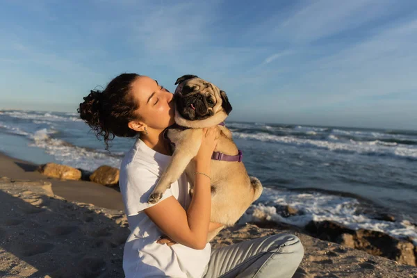 Junge Frau mit lockigem Haar küsst Mops Hund am Strand in der Nähe des Meeres in Barcelona — Stockfoto