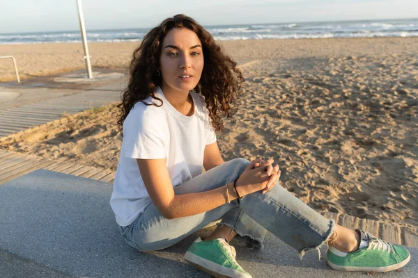 Bonita mujer con el pelo rizado sentado en la playa de arena cerca del mar en Barcelona - foto de stock