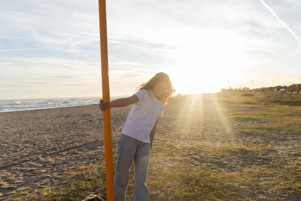 Positive young woman with curly hair standing near yellow pole against sun on beach in Barcelona — Stock Photo