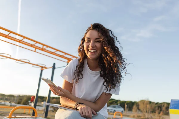 Vista de ángulo bajo del freelancer positivo sosteniendo teléfono inteligente cerca de la computadora portátil en la playa en España - foto de stock