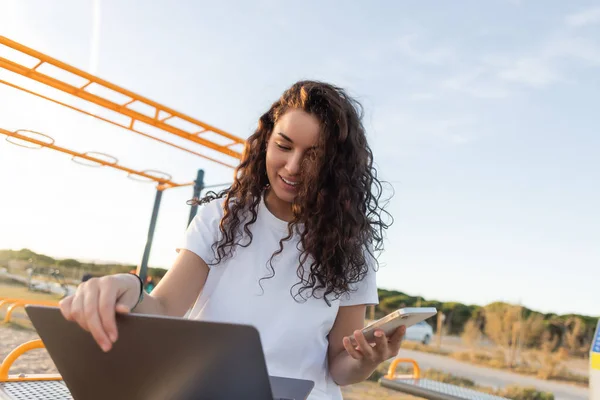 Alegre freelancer sosteniendo teléfono inteligente mientras usa el ordenador portátil en la playa en España - foto de stock