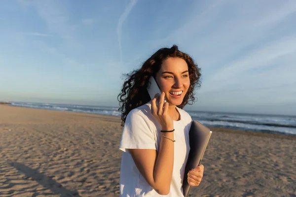 Cheerful freelancer holding laptop while talking on smartphone on beach in Barcelona — Foto stock