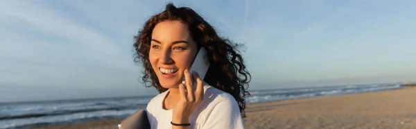 Alegre freelancer sosteniendo portátil mientras habla en el teléfono inteligente en la playa en España, bandera - foto de stock