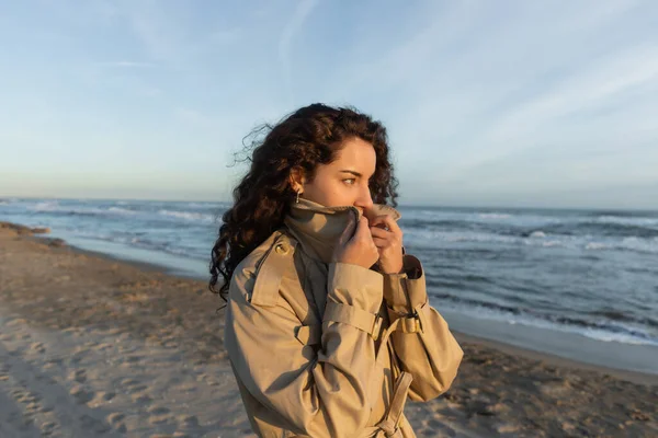 Curly young woman covering face with collar of beige trench coat on beach in Barcelona — Stock Photo