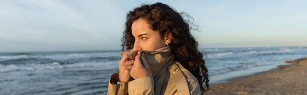 Curly young woman covering face with collar of beige trench coat on beach in Barcelona, banner — Stock Photo