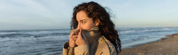 Curly young woman covering face with collar of beige trench coat on beach in Barcelona, banner — Photo de stock