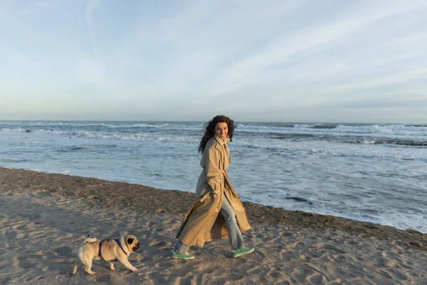 Full length of cheerful woman in trench coat walking with pug dog near sea in Barcelona — Stockfoto