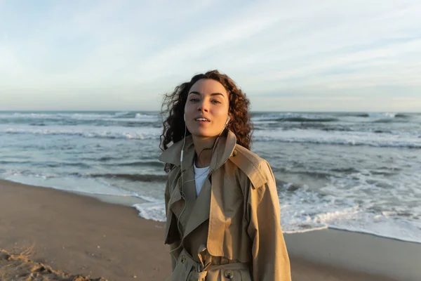 Curly young woman in beige trench coat and wired earphones listening music near sea in Barcelona — Photo de stock