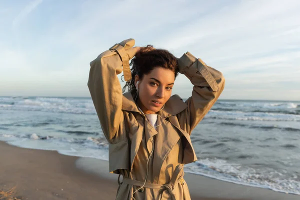 Mujer joven en gabardina beige y auriculares con cable escuchando música mientras ajusta el pelo cerca del mar en Barcelona - foto de stock