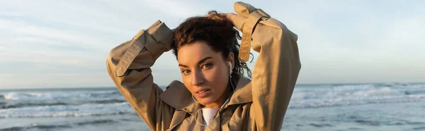 Giovane donna in trench beige e auricolari cablati ascoltare musica mentre regola i capelli vicino al mare a Barcellona, banner — Foto stock