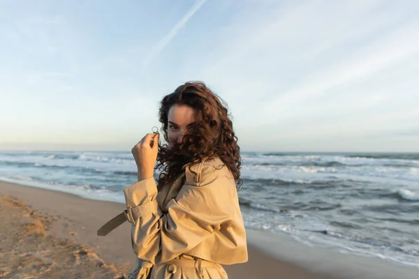 Young woman in beige trench coat adjusting curly hair while looking at camera near sea in Barcelona — стокове фото