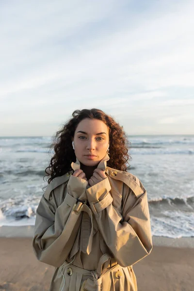 Curly young woman in wired earphones listening music while adjusting collar of beige trench coat near sea in Barcelona — Stock Photo
