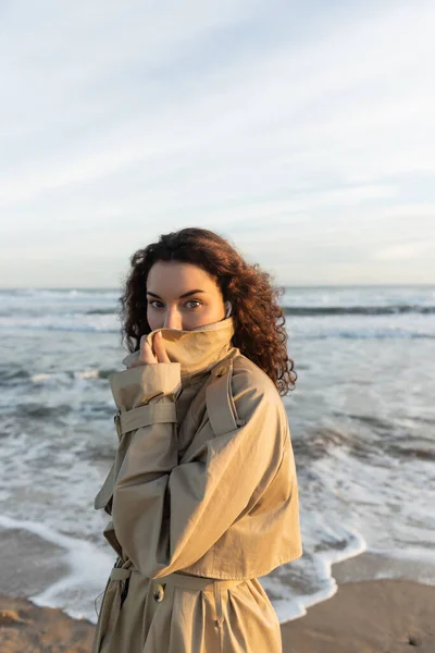Curly woman in wired earphones adjusting collar of beige trench coat and looking at camera near sea in Barcelona — стокове фото