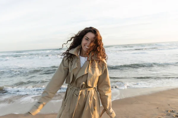 Cheerful young woman in beige trench coat and wired earphones listening music near sea in Barcelona — стокове фото