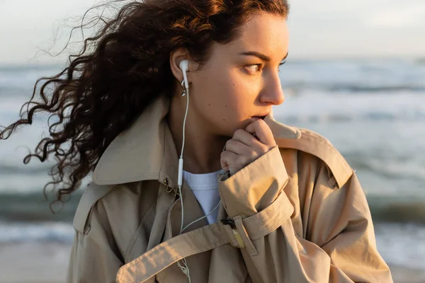 Porträt einer lockigen jungen Frau in beigem Trenchcoat und kabelgebundenen Kopfhörern, die am Meer in Barcelona Musik hört — Stockfoto