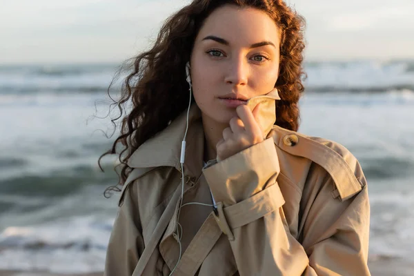 Portrait of curly woman in beige trench coat and wired earphones listening music near sea in Barcelona — Stockfoto