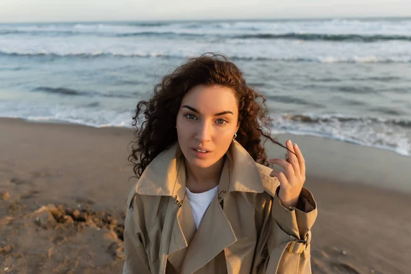 Curly woman in trench coat looking at camera on blurred beach near sea — Stock Photo