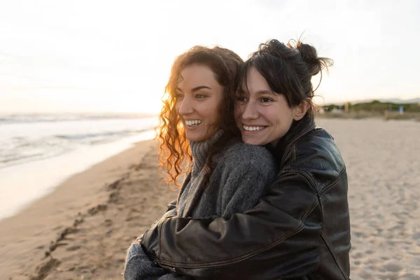 Улыбающаяся женщина обнимает друга на размытом пляже на закате — стоковое фото