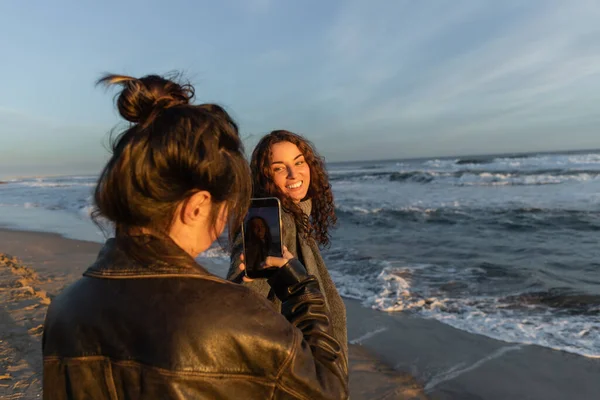 Woman taking photo on smartphone of smiling friend on beach in Barcelona — Stock Photo