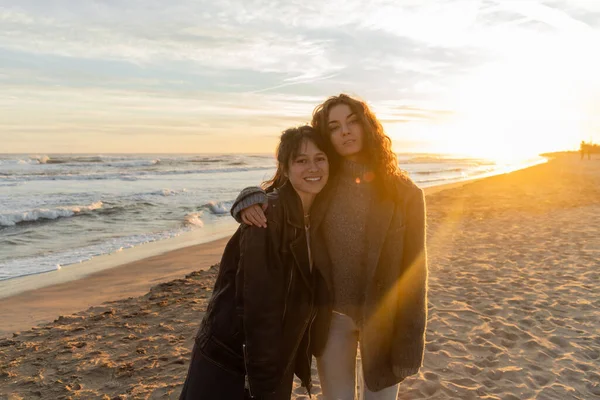 Curly woman hugging smiling friend on beach during sunset in Barcelona — Stock Photo