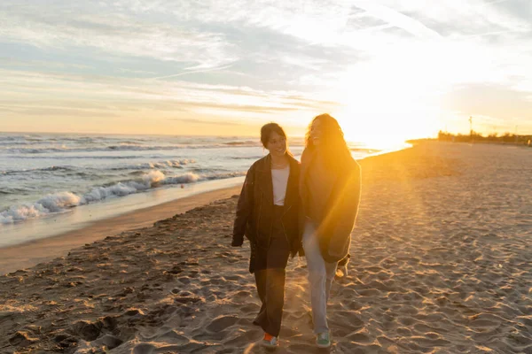 Femmes positives marchant sur la plage près de la mer pendant le coucher du soleil — Stock Photo