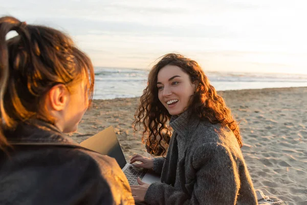 Sonriente mujer rizada usando el ordenador portátil y hablando o amigo en la playa - foto de stock