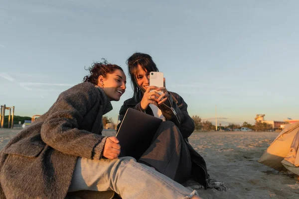 Amigos sonrientes usando smartphone y laptop en la playa en España - foto de stock