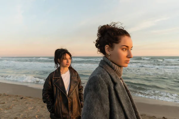 Mujeres jóvenes mirando hacia otro lado mientras están de pie en la playa en España - foto de stock