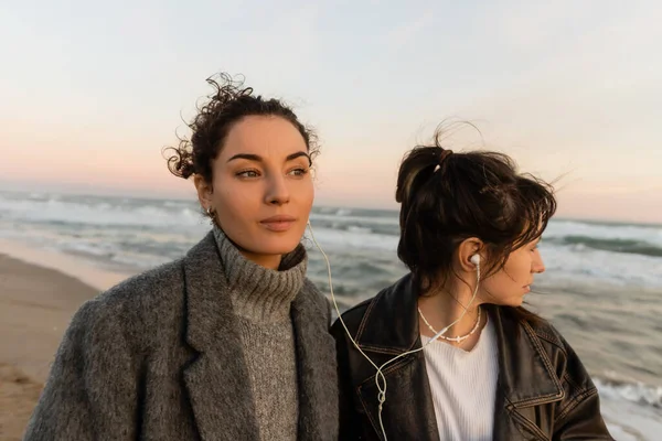 Curly woman listening music in wired earphones with friend on beach in Barcelona — Stock Photo