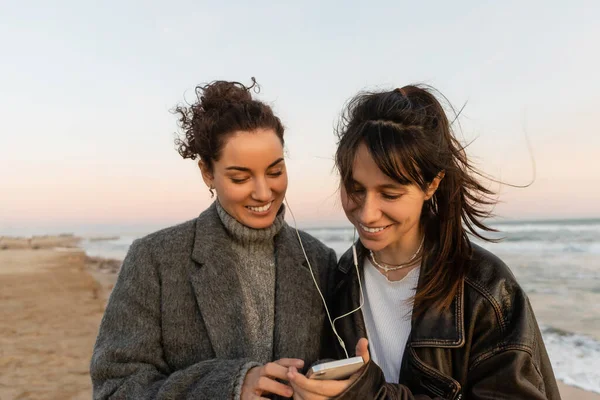 Positive friends in wired earphones using cellphone on beach in Spain — Foto stock