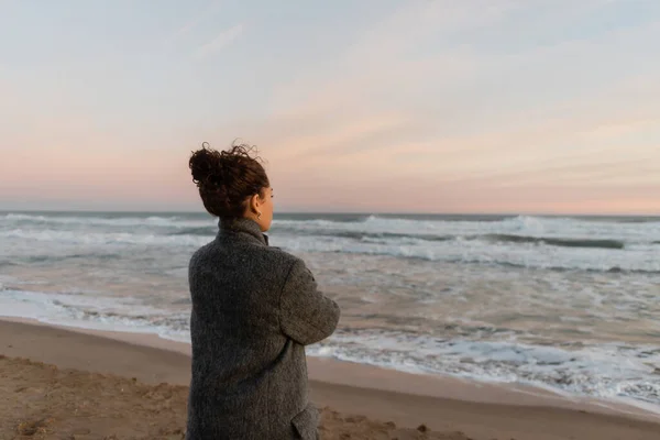 Кудрявая женщина в пальто смотрит на море, стоя на пляже в Барселоне — стоковое фото