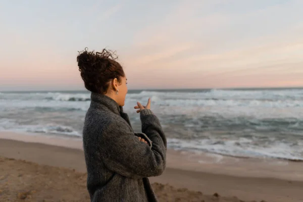 Lockige Frau im Mantel zeigt am Strand auf das Meer — Stockfoto