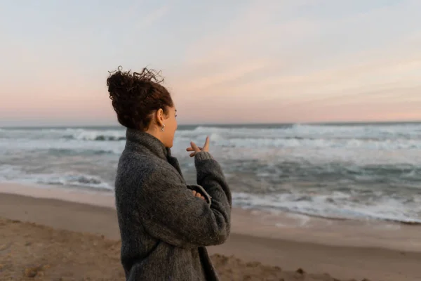 Curly woman in coat pointing at sea while standing on beach — Stock Photo