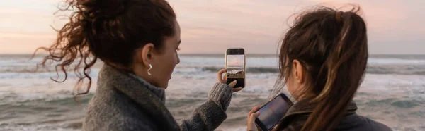 Friends taking photo on smartphones near sea during sunset, banner — Foto stock