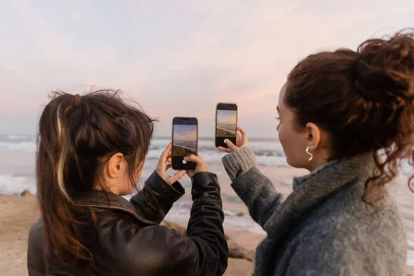 Young friends taking photo on smartphones while standing on beach in Spain — Foto stock