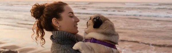 Side view of cheerful young woman looking at pug dog near sea in Spain, banner — Photo de stock
