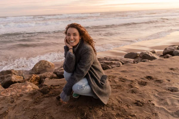Mujer positiva en abrigo mirando la cámara cerca de piedras en la playa de arena cerca del mar - foto de stock