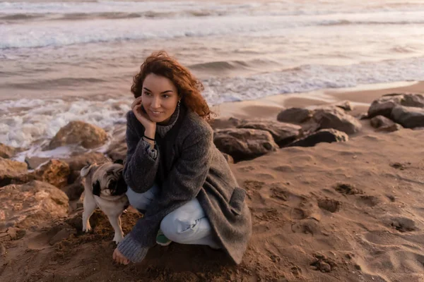 Mulher sorridente de casaco olhando para longe perto de cão pug na praia em Barcelona — Fotografia de Stock