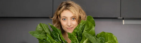 Пронзительная молодая женщина улыбается и держит зеленые листья капусты на кухне, баннер — стоковое фото