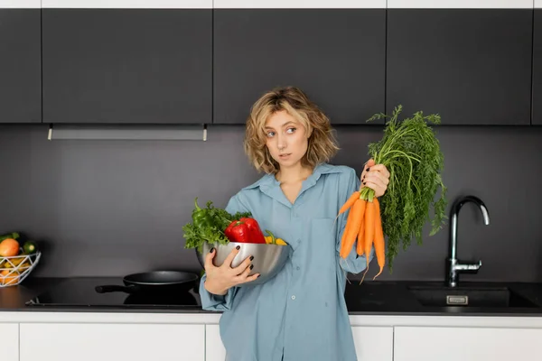 Смущенная молодая женщина держит миску с овощами и свежей морковью на кухне — стоковое фото