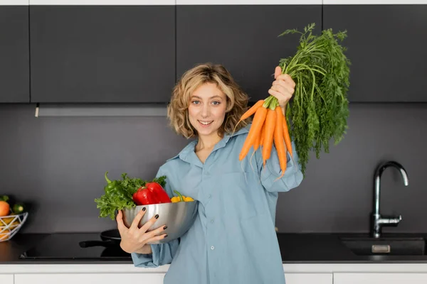 Cheerful young woman holding bowl with vegetables and fresh carrots in kitchen — Stock Photo