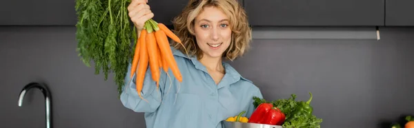 Веселая молодая женщина держит миску с овощами и свежей морковью на кухне, баннер — стоковое фото