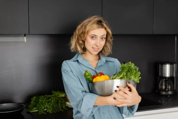 Cheerful young woman holding bowl with fresh vegetables in kitchen — Photo de stock