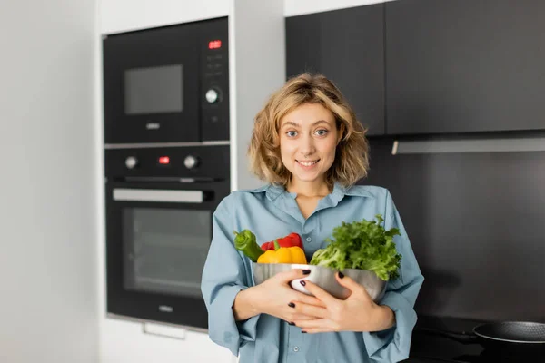 Positive jeune femme tenant bol avec des légumes frais dans la cuisine — Photo de stock