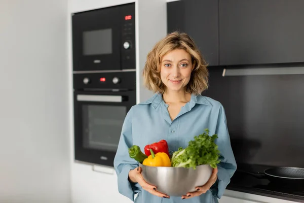 Довольная молодая женщина держит миску со свежими овощами на кухне — стоковое фото