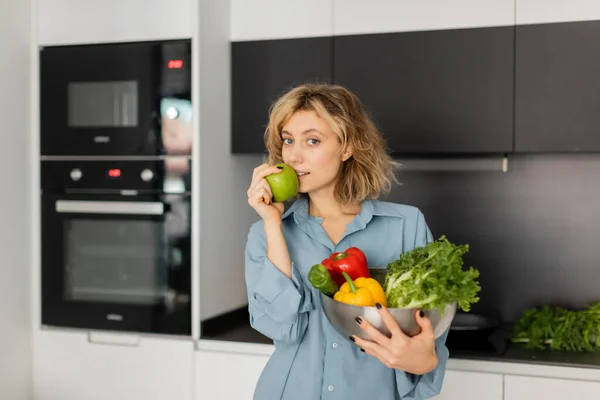 Blonde jeune femme tenant bol avec des légumes frais et pomme verte dans la cuisine — Photo de stock