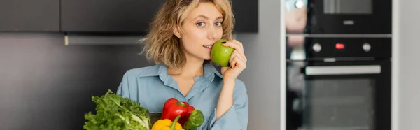 Blonde jeune femme tenant bol avec des légumes frais et pomme verte dans la cuisine, bannière — Photo de stock