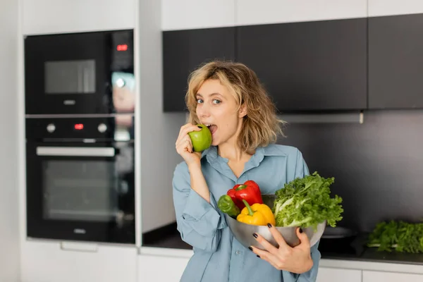 Femme blonde mordant pomme et tenant bol avec des légumes frais dans la cuisine — Photo de stock